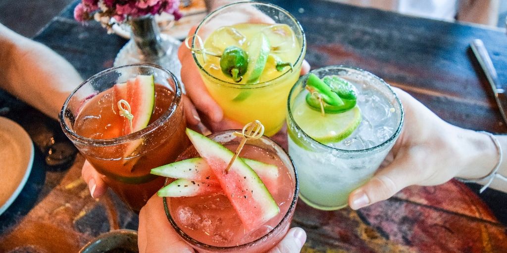 Cheers! Four people are holding margarita glasses together. Two are topped with sliced watermelon, and two of the glasses are topped with lime and Jalapeño slice.